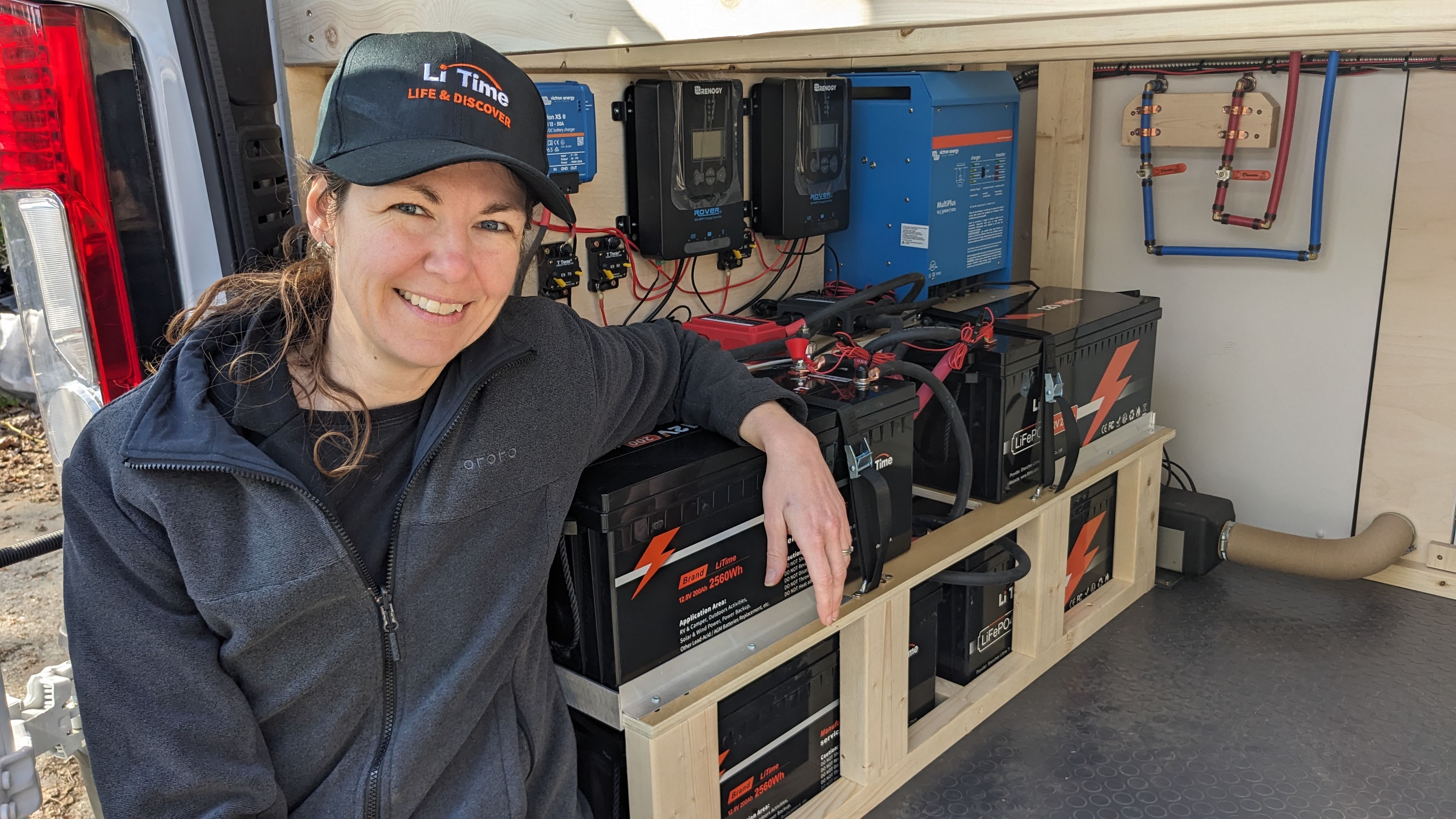 A woman in a Li Time cap leans smiling against her large, custom battery bank inside her van, featuring multiple LiTime batteries and inverters
